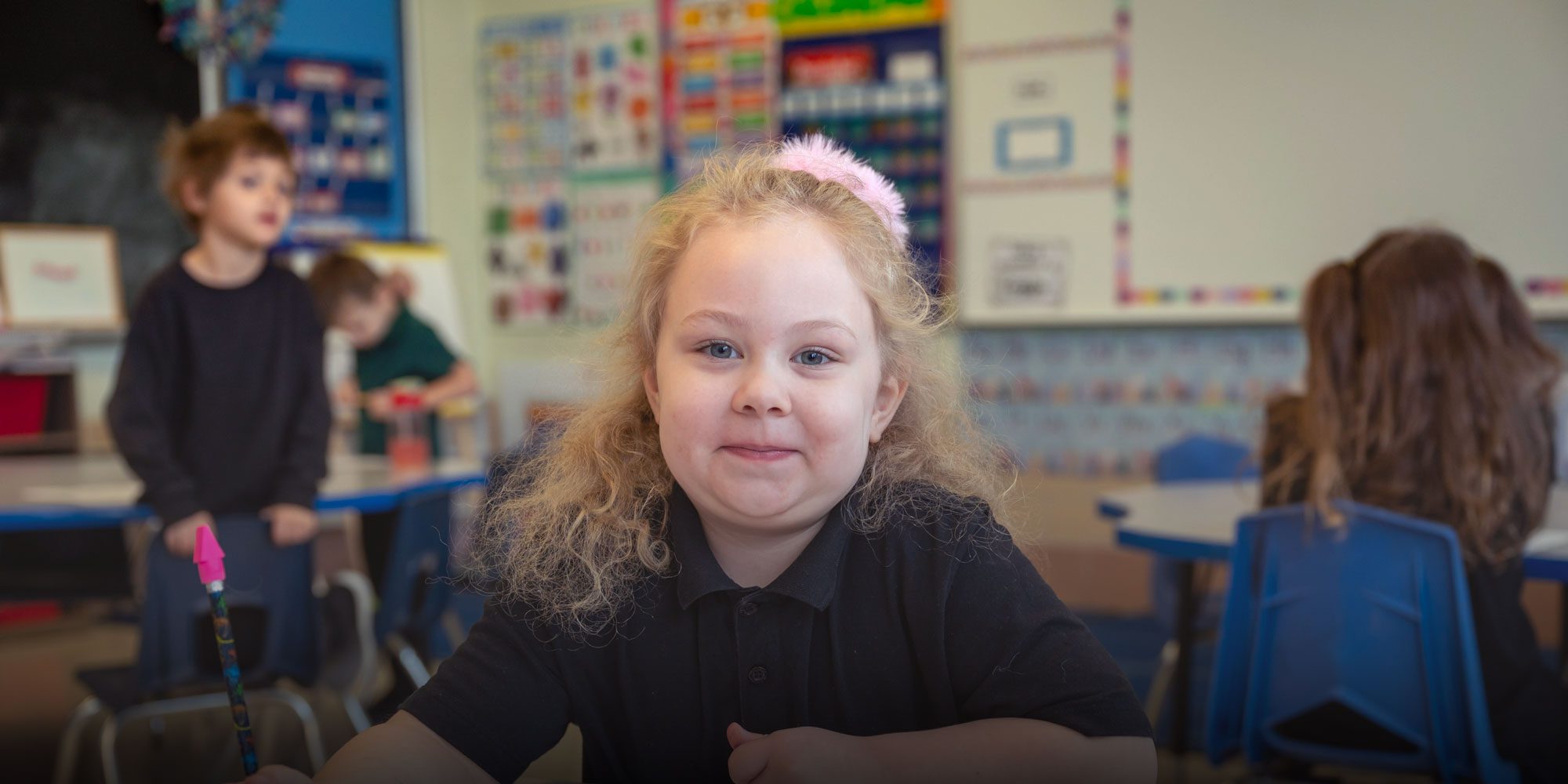 Smiling student with a pink headband sitting at a classroom desk.